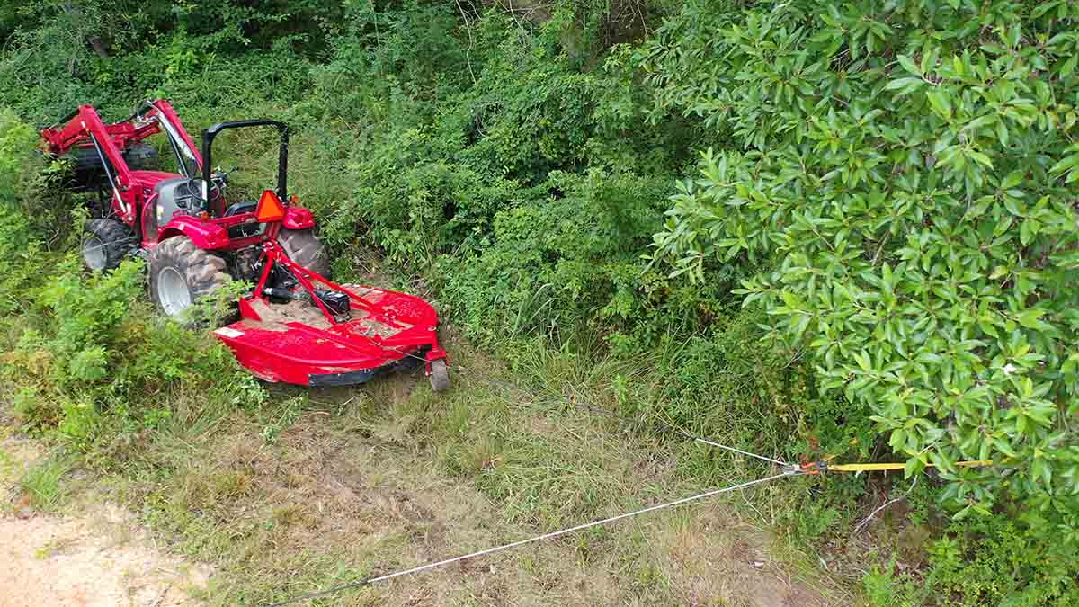 four wheel drive tractor with brush cutter, also called a bush hog, stuck in the edge of a swamp along a dirt road in the summertime being recovered with a winch using synthetic rope instead of wire